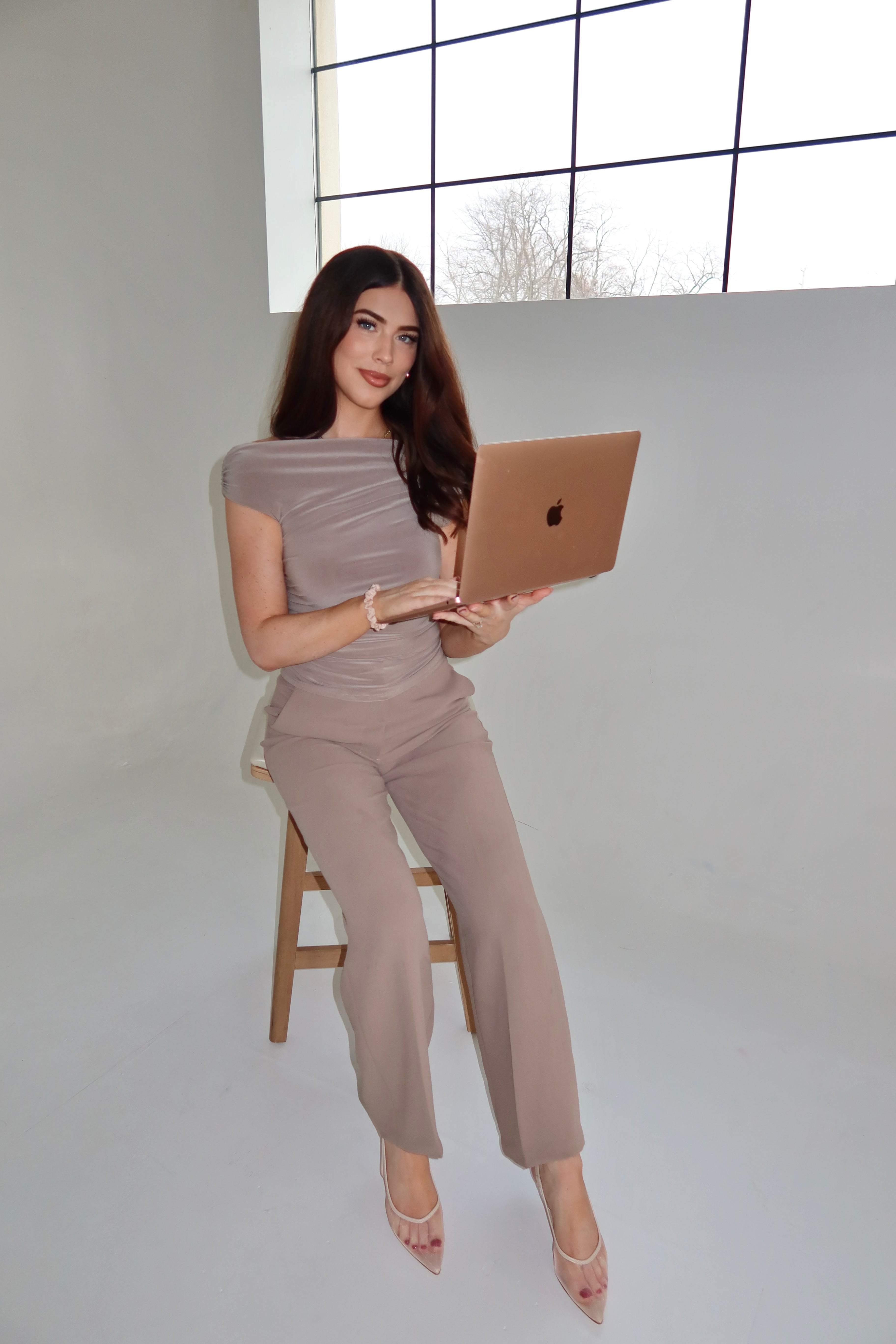 Woman sitting on a chair holding a laptop in a minimalistic room with large windows.